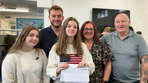  A young woman, a young man, and a middle-aged man and woman. They are all smiling at the camera and Megan is holding her results, on a sheet of paper, in front of her.