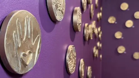 Zoie O'Brien/BBC A collection of gold coins put out on a table with a purple table cloth. The round coin closest to the camera has a hand with a heart inside the hand.