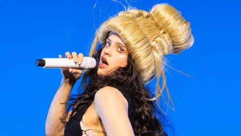 Samir Hussein Roslaia on stage wearing a blonde beehive wig and singing into a handheld microphone