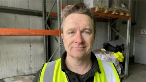 Jim Whiddett from Kent Trading Standards is standing in a building at a port, he is wearing a high vis jacket.