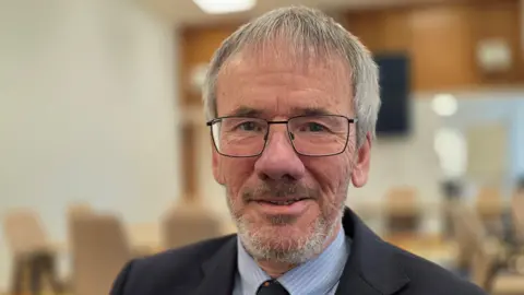 Paul Moseley/BBC Brian Watkins is wearing glasses. He has a navy suit jacket over a blue and white striped shirt with a tie.