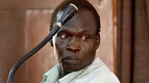 Getty Images Thomas Kwoyelo looks into the distance during a war crime trial in Uganda. He wears a mint green shirt and sits behind a microphone.