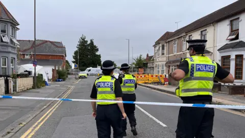 Three police officers stood near police tape inside of a cordon on Manchester Road. They are wearing yellow high vis jacket saying 'police'. There are several houses in the background. 