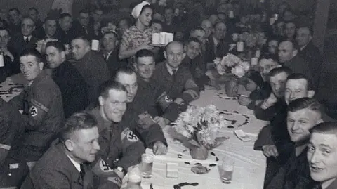 Uniformed German soliders with swastika arm bands sit around long tables drinking from glasses and mugs