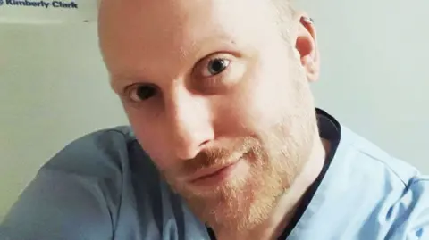 Close-up of a white blonde man in his 30s, with a light beard and wearing blue scrubs, as he smiles at the camera.