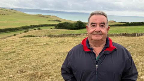 A farmer in a blue coat with red collar stands in a field close to the coast. Rolling fields are in the background and beyond that the sea. 