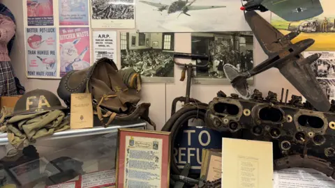 A World War Two display at Midlands Air Museum includes documents, uniforms, a mechanical component and a model aircraft. There are World War Two posters on the wall.