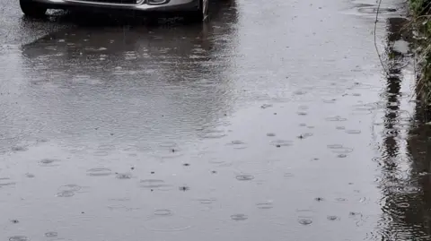 Bob Ashworth An image of a flooded pavement, with the wheels of a car in the background, and rain spots on the standing water.