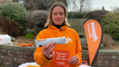 Katie White stands outside, looking to camera and holding up a self-swab DNA kit, which is inside a white bag with the word Enough on it. She is also holding a flyer that says 'Together we can end rape'. She is wearing a bright orange hoody. 