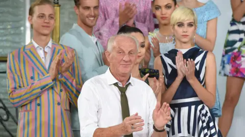 Getty Images Paul Costello wears a white shirt and green tie. Behind him are female and male models wearing bright clothes.