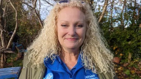 Maria Perrett looking at the camera smiling. She has long blonde curly hair and is wearing a blue three-quarter zip jumper and a khaki raincoat. 