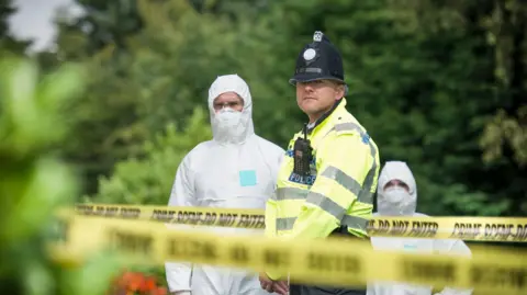 Monty Rakusen/Getty Police officer standing by crime scene tape with forensic investigators in white overalls and trees behind him. 