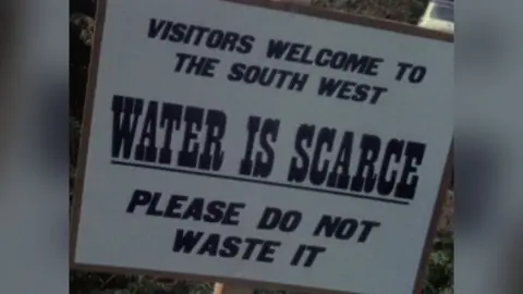 A white sign is attached to a wooden post. In bold letters it reads: "Visitors Welcome to the South West, Water is scarce. Please do not waste it."