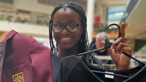 Emma Baugh/BBC A teenager is grinning at the camera and holding two clothes hangers with a blazer and a pullover. She has long plaited hair and dark-rimmed spectacles.