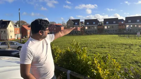 Eric Johnson/BBC A man in a white T-shirt stands outside his home on a sunny day pointing towards a new housing estate.