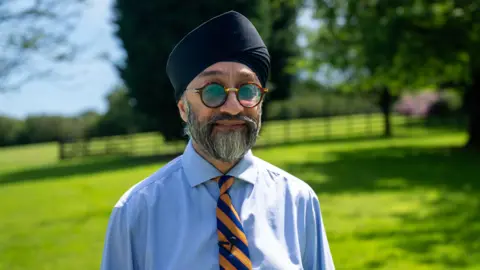 A man stands in his garden, smiling and looking at the camera, wearing a turban and the striped tie of his great grandfather's War regiment
