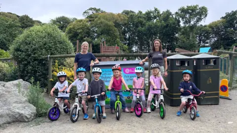 A row of children about age four and five on bikes, two women standing behind, at a playground. Trees and blue skies seen behind.