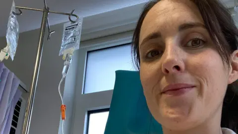 Sammy is sitting in a hospital chair. She is wearing a light grey jumper with a zip on it. She has dark brown hair which is tied back off her face. There is a drip bag connected to a stand in the background of the picture which is attached to Sammy.
