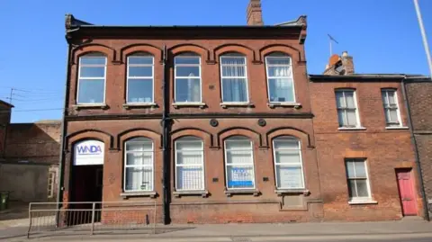 BELTON DUFFEY The outside of a brick building with nine large windows. Over the door is a sign in purple letters saying WNDA standing West Norfolk Deaf Association