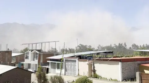 A haze of dust rises from a settlement with one and two-storey buildings. There are mountains in the background. 