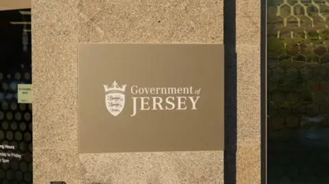 BBC A close-up of the Government of Jersey sign attached to a concrete pillar. On the sign, white text is printed on a brown background.