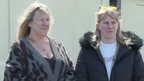 Two women with blonde hair stand side by side. One wears a black puffer jacket the other a leopard print cardigan.
