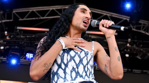 Getty Images Shot of singer Leo Kalyan on stage at the Trafalgar Square Show on Saturday 4 July. He wears a white and black strappy top and sings into the mike. He has long dark hair and light beard growth.