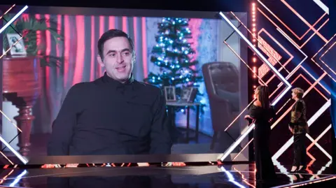 Ronnie O'Sullivan on screen at the BBC Sports Personality of the Year awards in 2020. He is projected on a big screen while sat in front of a Christmas tree and wearing a black jumper. The presenters on stage are looking at him on screen.