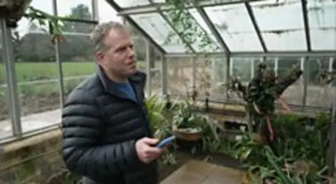 A man in a padded winter coat is in a large greenhouse holding a phone in his right hand. He is listening to a plant in front of him speak.