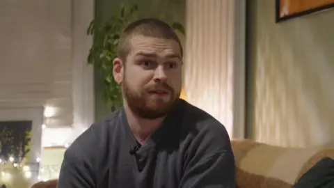 Conall Ó Brolcháin is seen sitting on a couch in his flat. Behind him are fairy lights and a house plant.