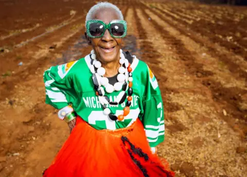 Luxury Media Zambia Margret Chola smiling in the field with her hand on her hip, wearing big jewel green sunglasses, a green sports jersey, a red dress and a big white, red and black plastic around her neck.