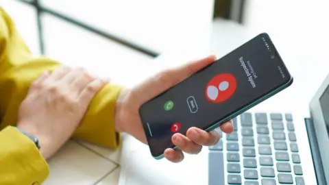 Getty Images A stock image of a woman looking at a smartphone which has a call coming it, it says "incoming call, suspected spam" on the phone.
