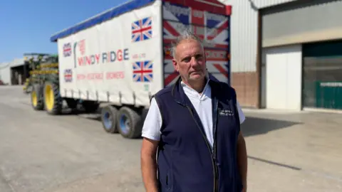 Mark Nundy - a man in a blue gillet - is standing in front of a tractor and trailer in a concreted farmyard