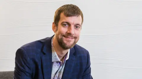 Jasco Foto de cabeza de Jason Trice sonriendo a la cámara. Tiene cabello castaño y barba castaña y viste camisa y chaqueta.