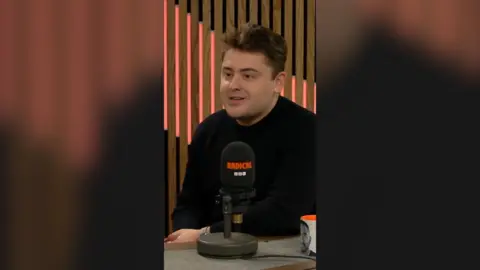 Joe Seddon has dark hair and is wearing a black long-sleeved top. He sits behind a mic