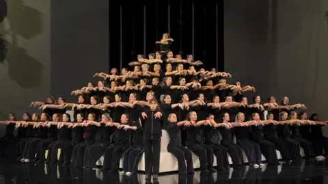 LMA University The dance group dressed all in black with outstretched arms. The stand in a pyramid formation, as part of a choreographed routine.