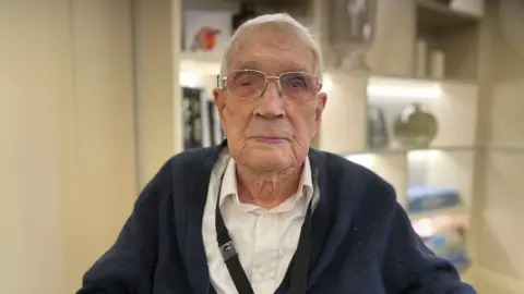 An elderly man wearing glasses and a blue cardigan looking straight at the camera. There's a set of shelves in soft focus behind him.