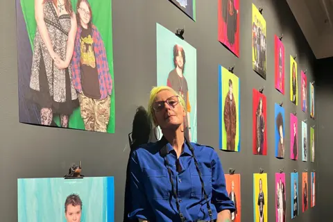 Casey Orr The photographer has short blonde hair and is wearing a blue shirt. She is standing in the exhibition space with more than a dozen of her photographs behind her. 