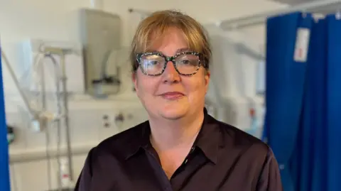Portrait of a Justine wearing patterned glasses and a dark shirt, standing in a clinical room with medical equipment and blue privacy curtains visible in the background.