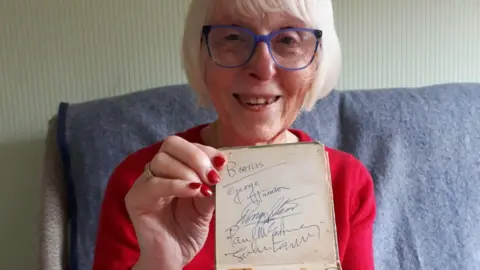 A woman in a red jumper with red painted nails holds up a notebook with Beatles signatures in it. She has white blonde hair and is wearing blue rimmed glasses. She is sitting on a sofa with a grey throw on it, with cream lined wallpaper behind her.