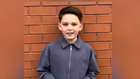 Family photo Child stands against a brick wall smiling he has dark hair and wears a grey jacket.