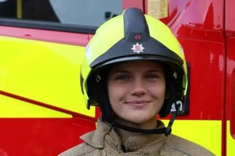 Essex County Fire & Rescue Service Phoebe Hutley is looking towards the camera smiling. She is wearing a yellow and black firefighter helmet that has an Essex County Fire & Rescue Service logo on the front. She is standing in front of a yellow and red fire engine.