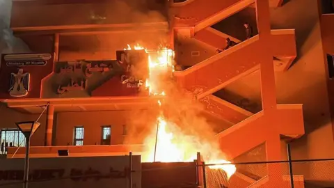 A fire on the outside staircase of Nasser Hospital throws up smoke as three people watch from higher steps