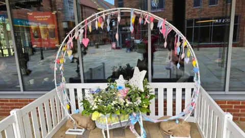 Kayleigh James A circular white display with colourful ribbons and hearts with a cot with green leaves, flowers and stars elevated on a white platform