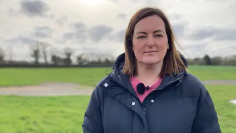 A woman wearing a navy coat and a pink jumper with a microphone attached to it. She has shoulder-length brown hair and is smiling. The background is blurred but a large green field and trees can be seen.