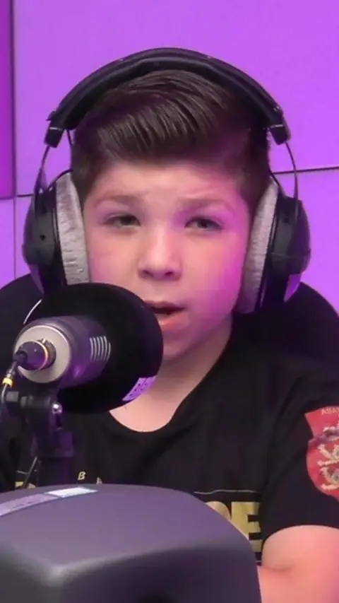 Boy talking into a microphone with headphones on and a purple background.
