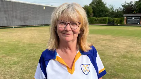 Ollie Conopo/BBC Linda Simms, looking straight at the camera and smiling, with shoulder length blonde hair, she is standing on a bowling green and is wearing a white top with blue and gold on it. A building is to her left, behind her.