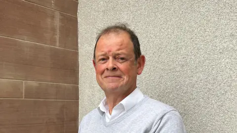 BBC/Marian McNamee Chris is wearing a light grey jumper over a white shirt. He is standing in front of a stucco-covered wall behind him and a red sandstone wall on the left of the picture.