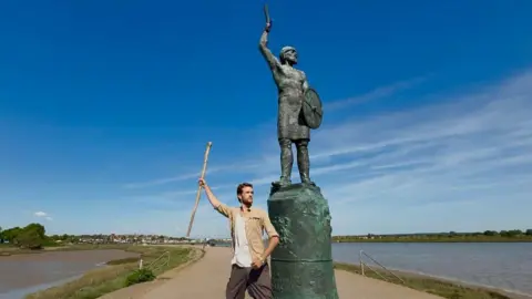 Amelie Mason/BBC James Lawrence poses holding his stick, mimicking the statue next to him. The water of Hythe Quay is visible left and right of him, with buildings in the background.