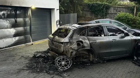 Bayliss family A burnt-out car with large pile of black debris on the ground with a detached wheel. There is a large garage door showing white heat marks and black scorching, and adjacent steps which are also scorched. There is some scorching on the block-paved drive.